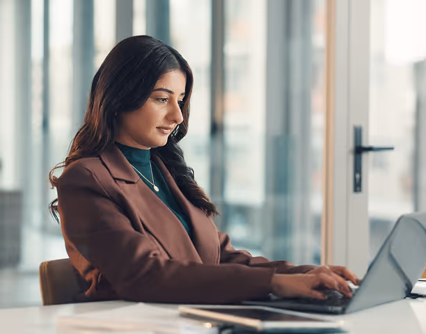 A woman wearing a brown blazer typing on a laptop at a desk in a bright office space.