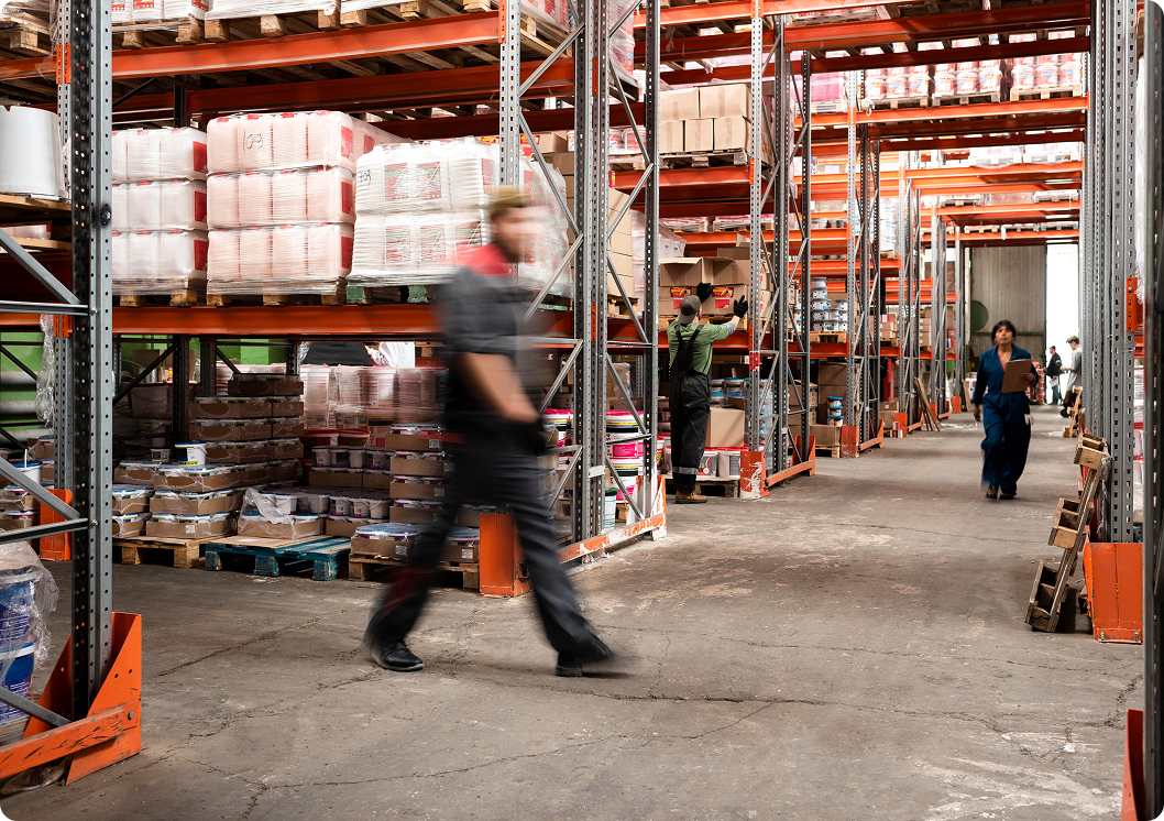 A female worker wearing a yellow high-visibility jacket, blue hard hat, and safety glasses inspects inventory in a warehouse. She holds a clipboard and checks labels on stacked metal pipes.