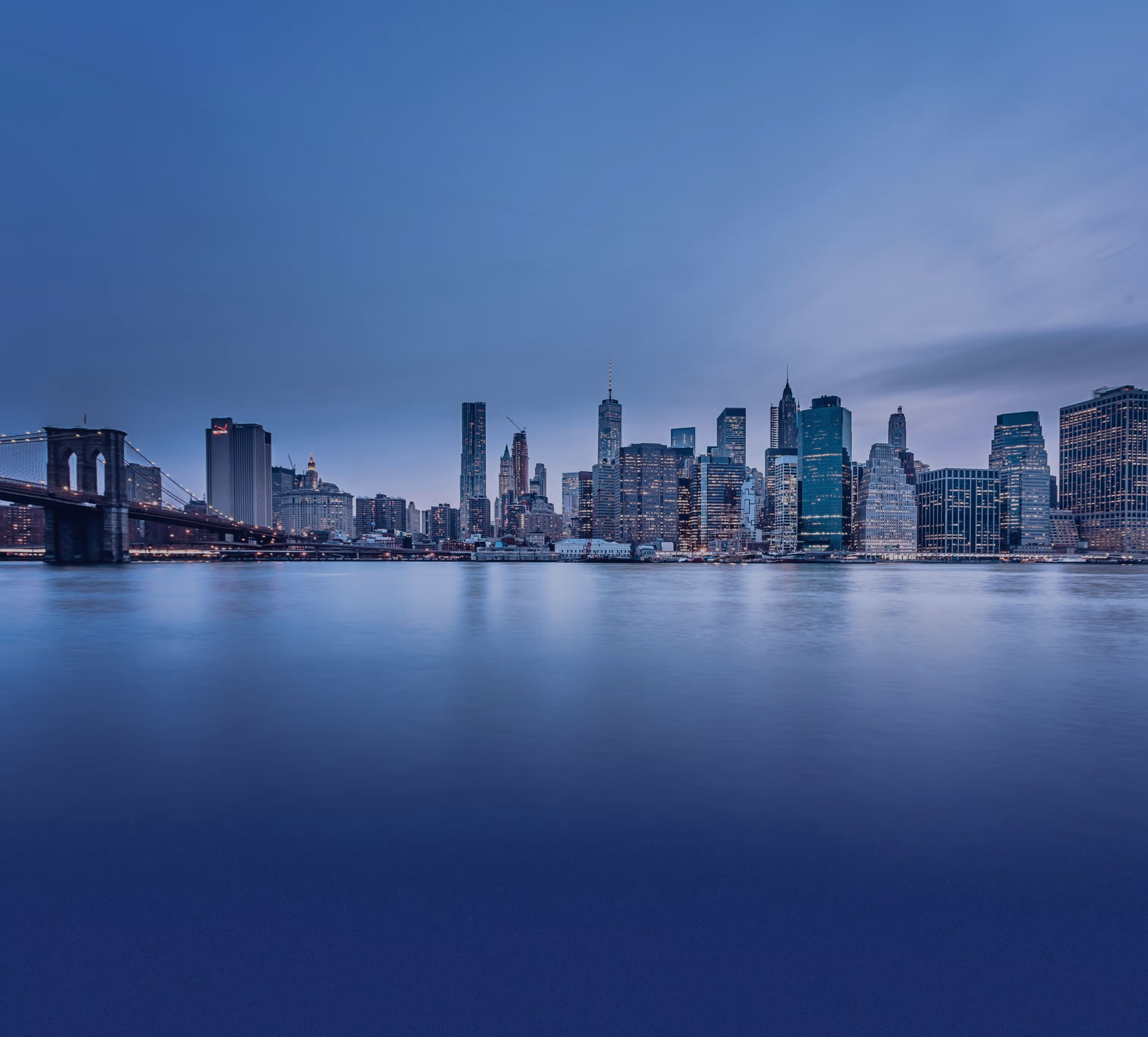Panoramic view of New York City skyline at dusk with illuminated buildings and Brooklyn Bridge over calm water.