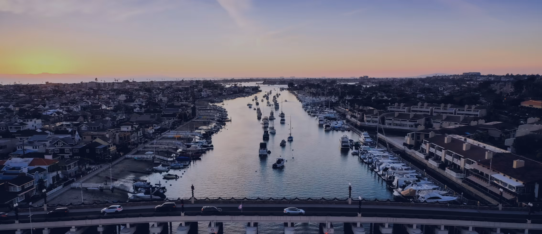 Aerial view of a marina canal at sunset with boats moored along both shores and a bridge crossing in the foreground.