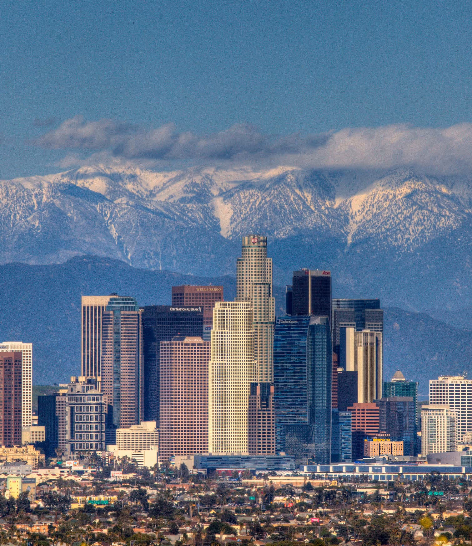 Downtown Los Angeles skyline with tall buildings in front of snow-capped mountains under a blue sky with some clouds.