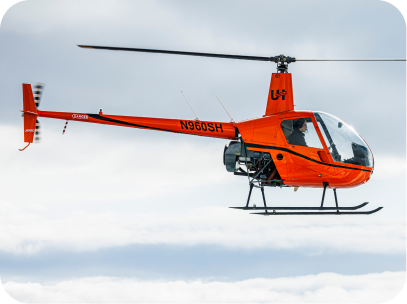 Orange Robinson R22 helicopter flying above clouds with pilot visible inside cockpit.