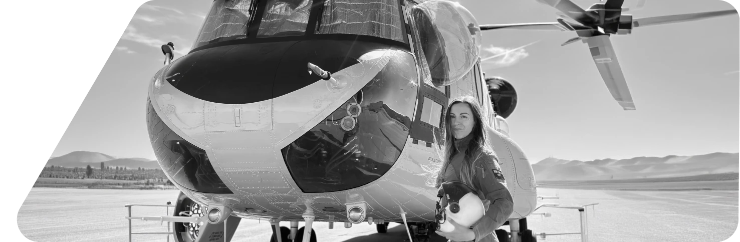 Woman in flight suit holding a helmet standing next to a helicopter on a runway with mountains in the background.