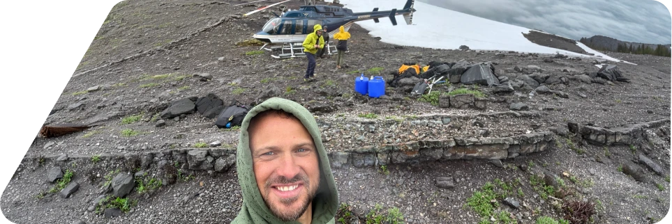 Man in a green hoodie taking a selfie on rocky terrain with two people near a helicopter in the background on a cloudy day.