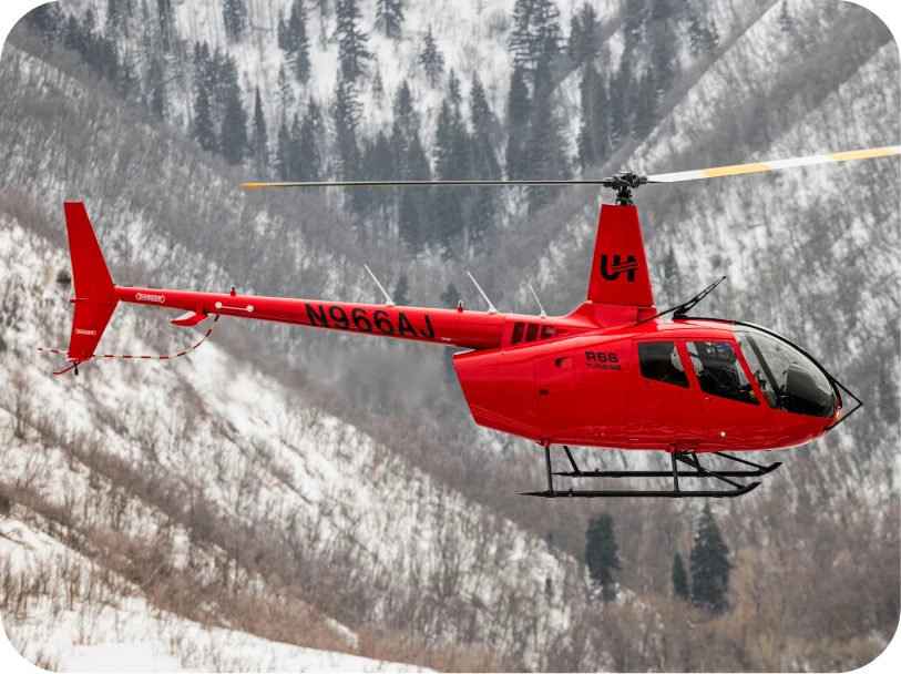 Red Robinson R66 Turbine helicopter flying over snow-covered mountains with trees.