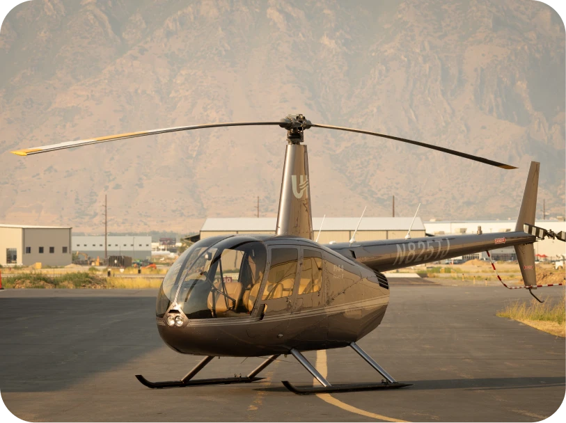 Gray Robinson R44 helicopter parked on an airport tarmac with mountains in the background.