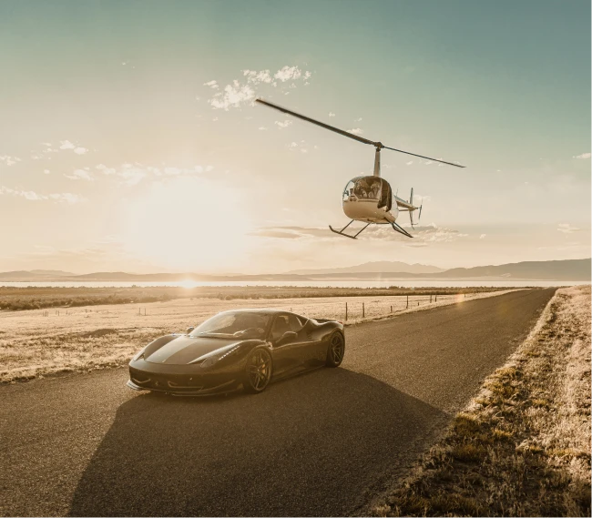 Black sports car driving on a rural road at sunset with a helicopter flying nearby.