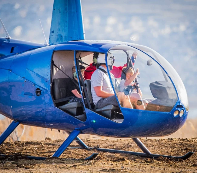 Blue helicopter on ground with four seated people inside wearing headsets.