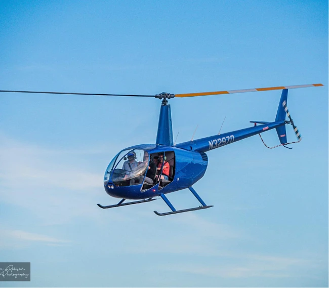 Blue helicopter flying in a clear sky with three visible occupants inside.