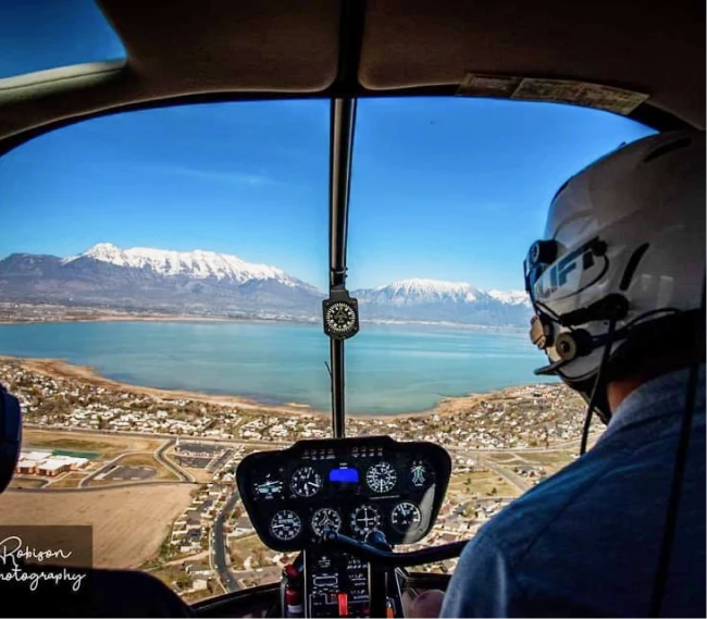 View from inside a helicopter cockpit with pilot wearing a helmet, overlooking a lake with snow-capped mountains in the background.