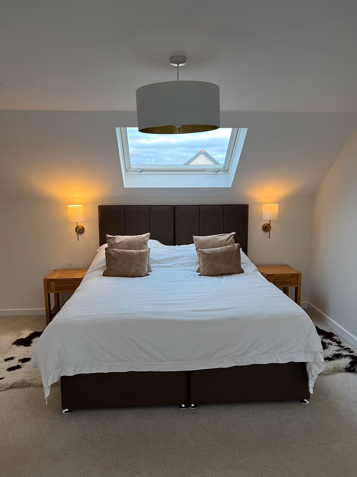 Modern bedroom with a large bed featuring a dark headboard, white bedding, four beige pillows, two wooden nightstands with lit wall lamps, a skylight window above the bed, and a ceiling light fixture.