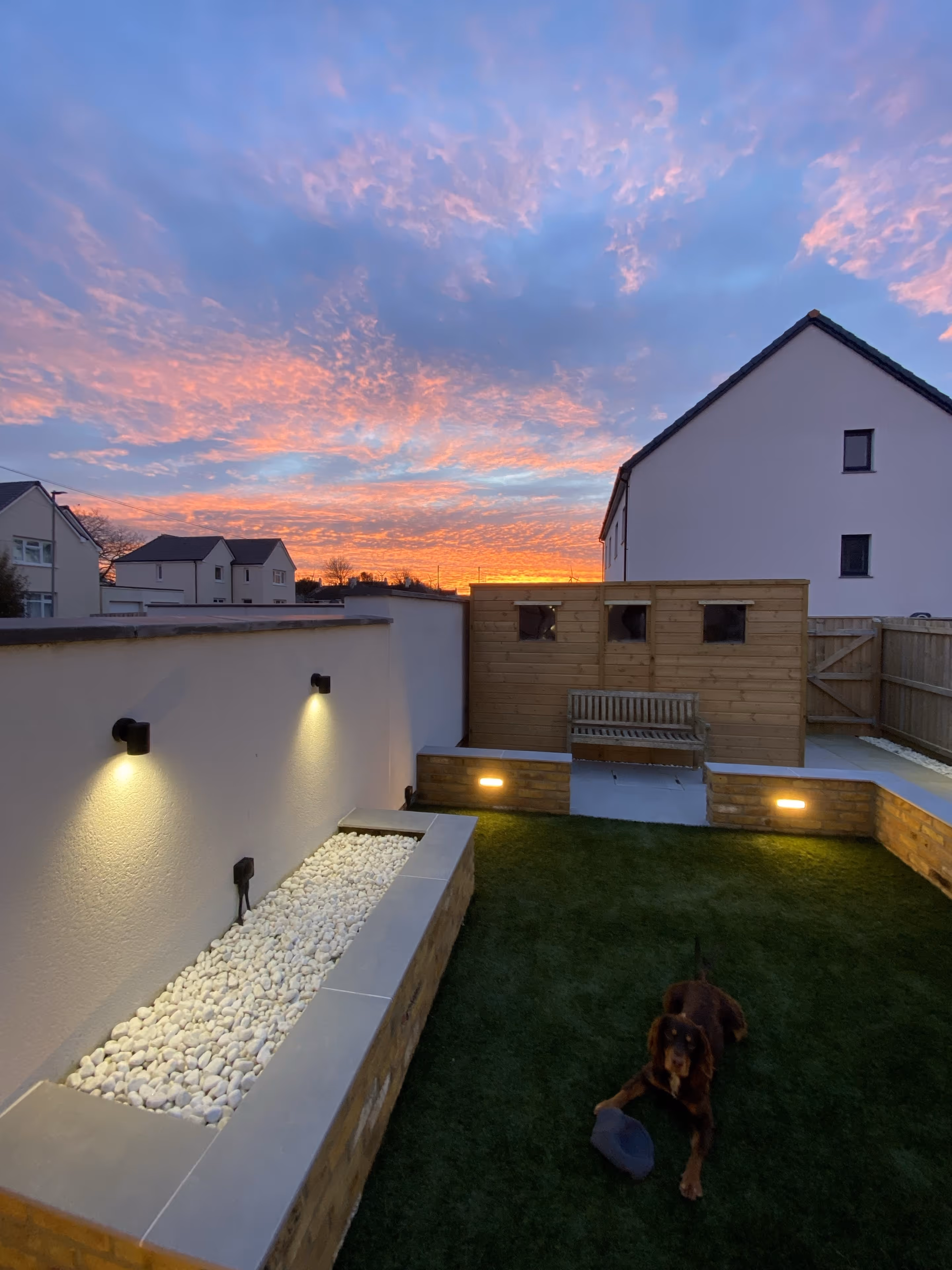 Backyard at sunset with lit wall lights, artificial grass, a wooden bench, and a brown dog lying near a toy.