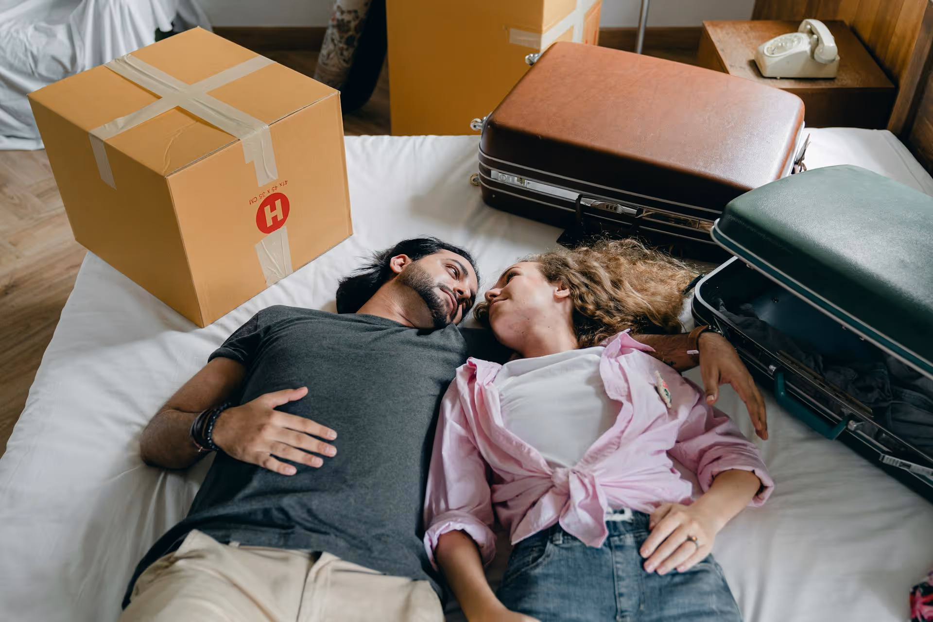 Couple lying on a bed surrounded by packed suitcases and a cardboard box, looking at each other.
