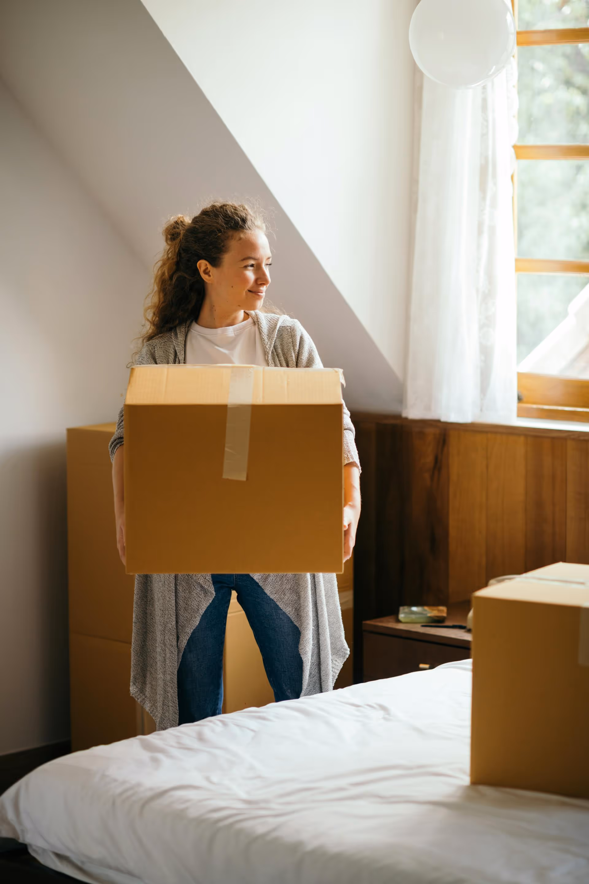Woman carrying a cardboard box in a bedroom with packing boxes around, next to a window with white curtains.