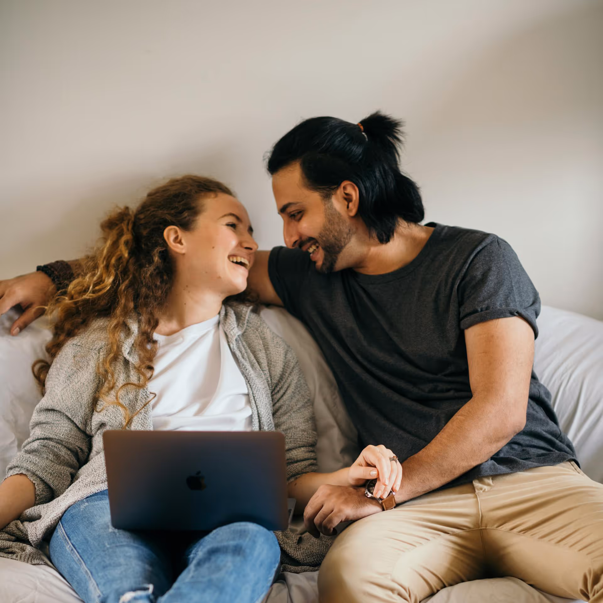 Couple sitting on a couch laughing and looking at each other with a laptop on the woman's lap.