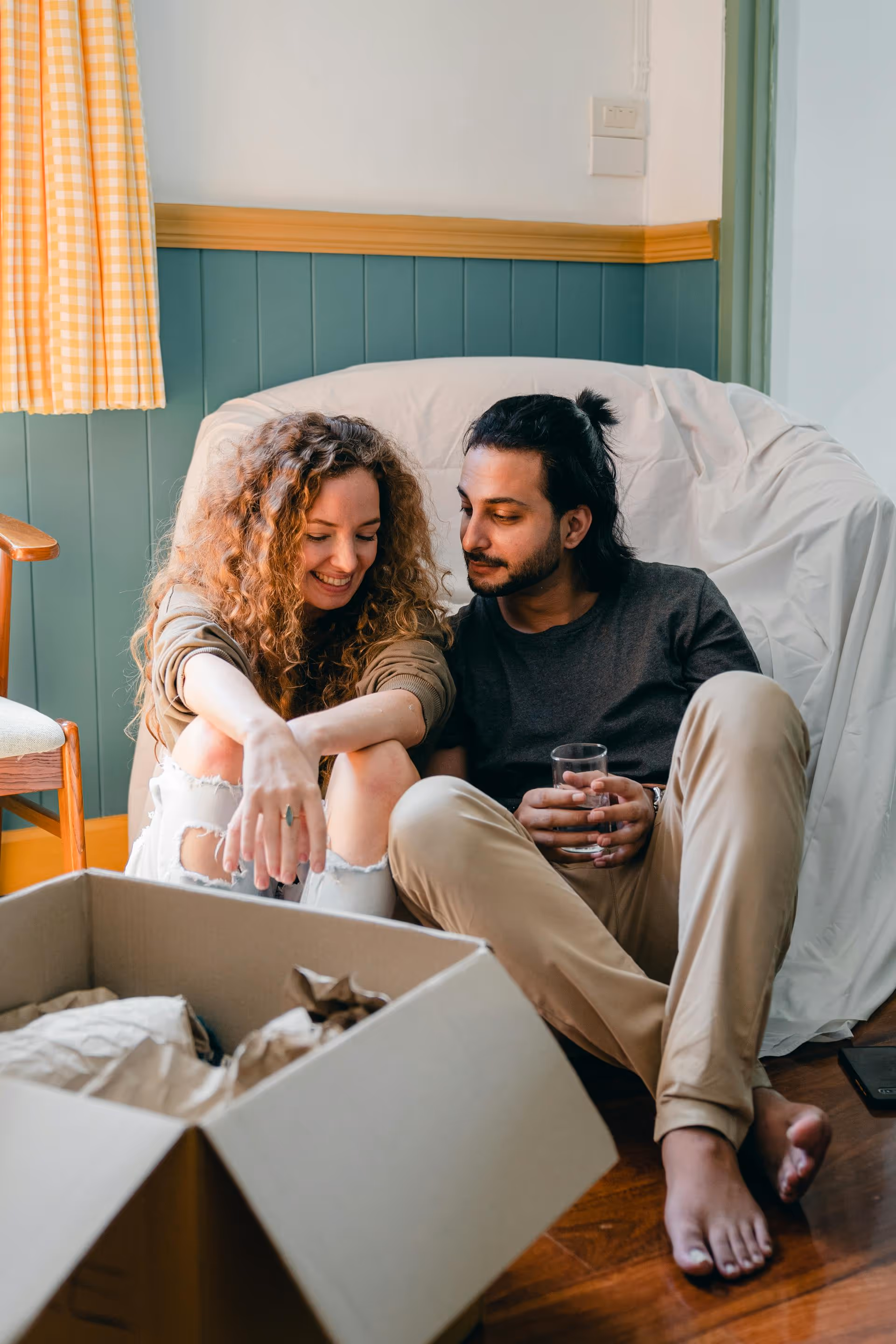 Young couple sitting on the floor by a white-covered chair, smiling and looking into a cardboard box.