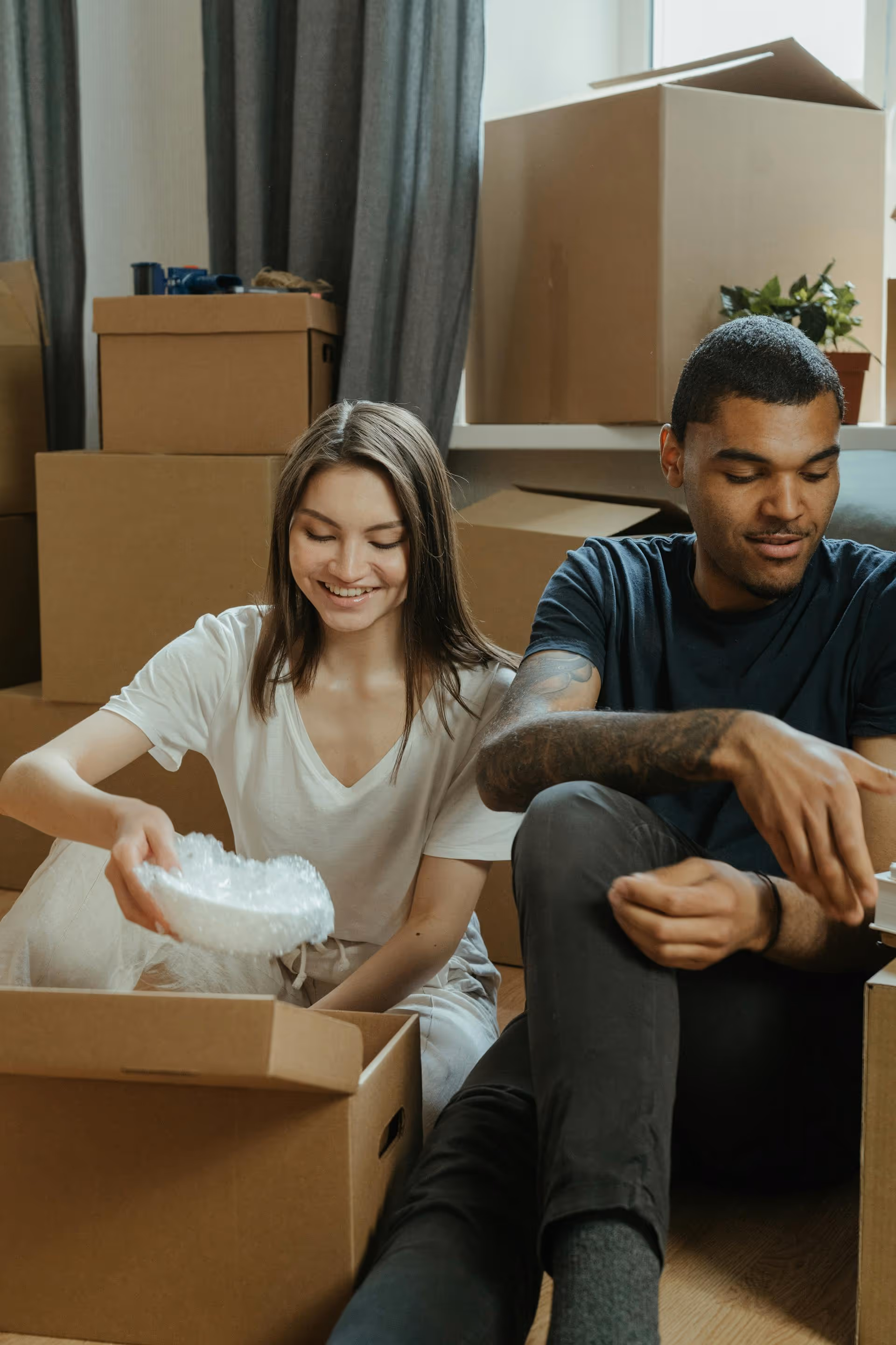 Smiling young woman and man packing items into cardboard boxes surrounded by stacked boxes in a room.