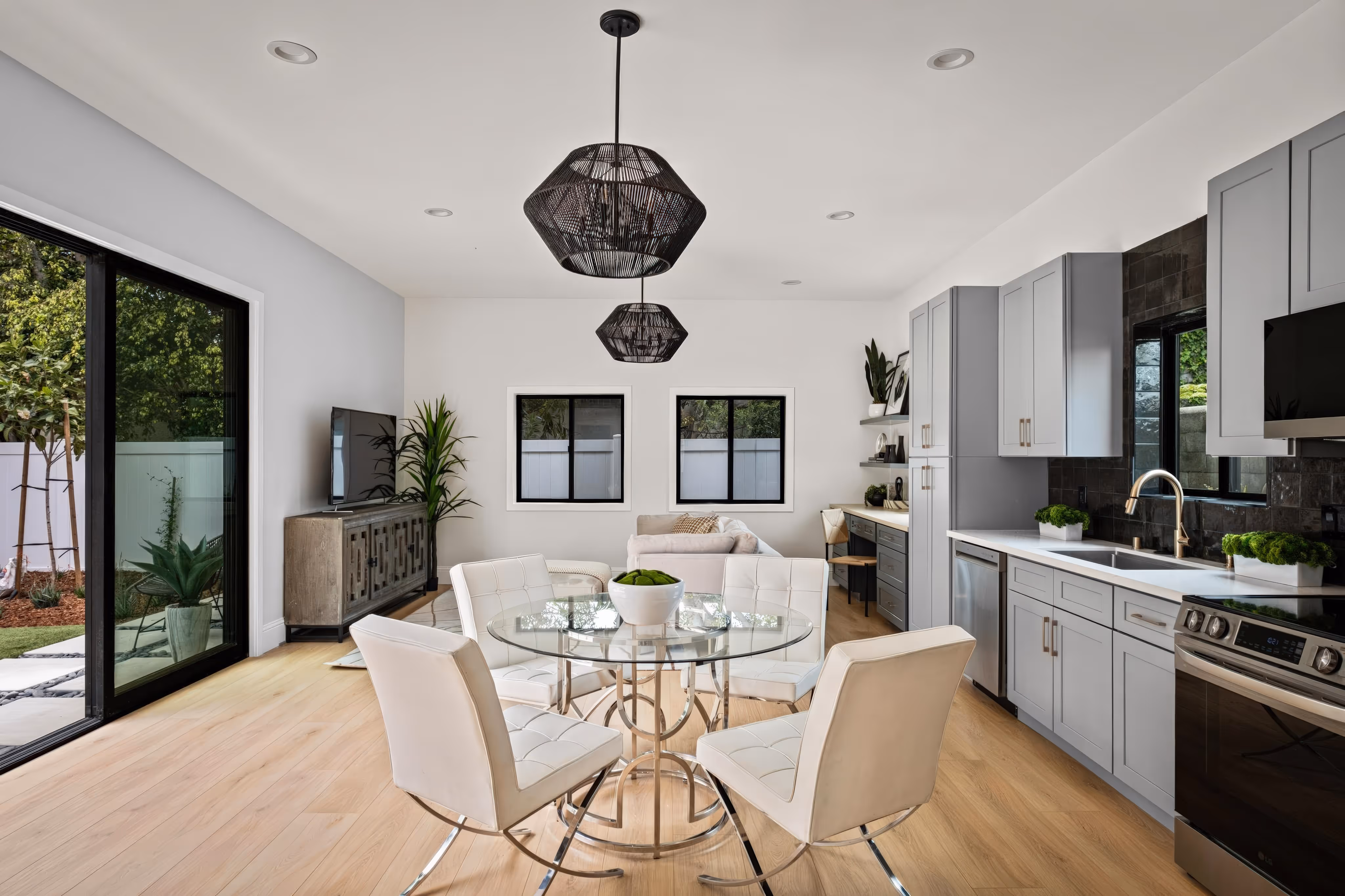 Modern open-plan kitchen and dining area with light wood floors, gray cabinets, glass dining table with white chairs, and sliding glass doors.