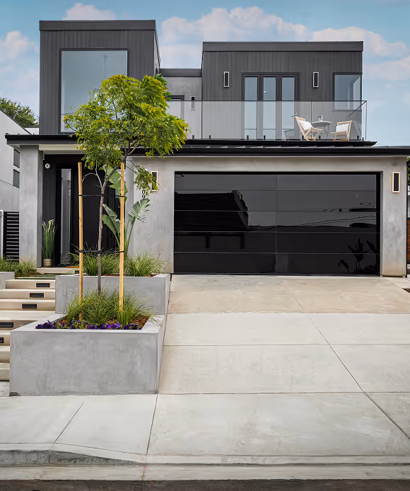 Modern two-story house with black garage door, balcony with chairs, and landscaping with small trees.