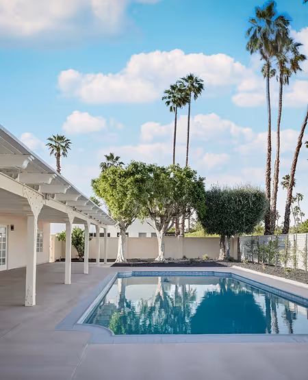 Outdoor swimming pool next to a white house with a covered patio and palm trees in the background under a partly cloudy blue sky.