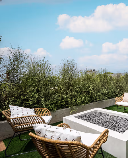 Outdoor seating area with rattan chairs and white cushions around a square fire pit filled with black stones, set against greenery and a blue sky with clouds.