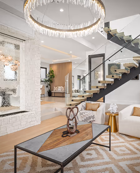 Modern living room with glass staircase, circular chandelier, white armchairs, and geometric patterned rug.