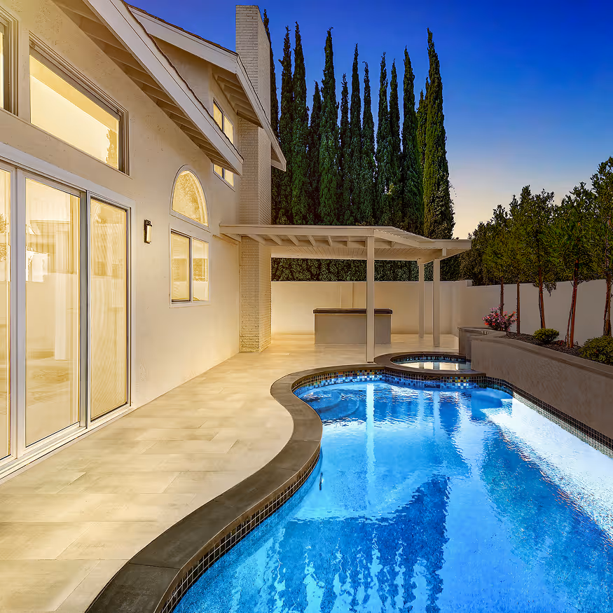 Modern backyard with curvy swimming pool, hot tub, covered patio, and tall trees in the background at twilight.