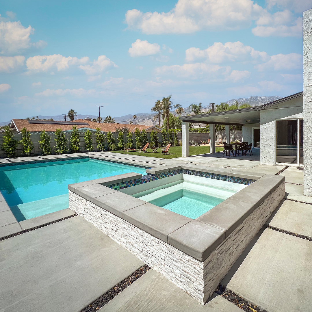 Modern backyard with a rectangular swimming pool, attached hot tub with stone trim, lawn with lounge chairs, and a covered patio with dining table.