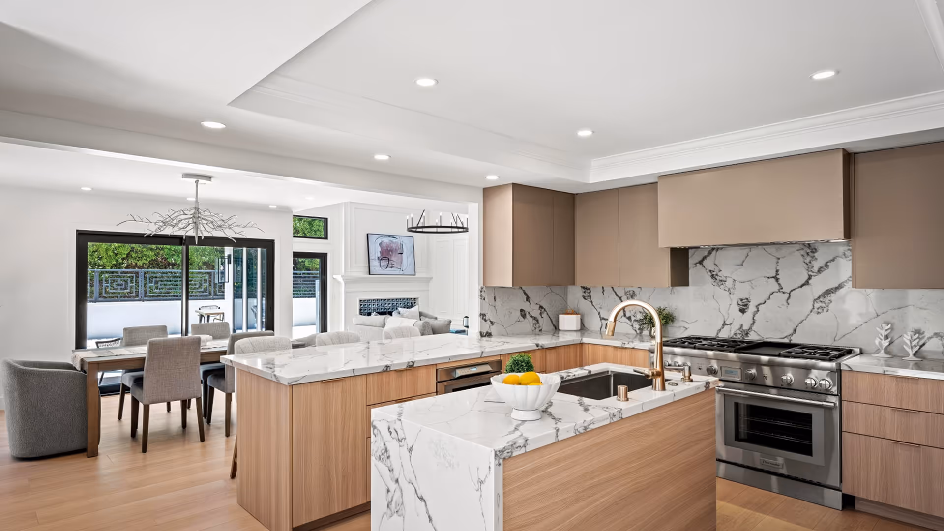 Modern kitchen with wood cabinetry, marble countertops, stainless steel stove, and dining area with large glass doors.