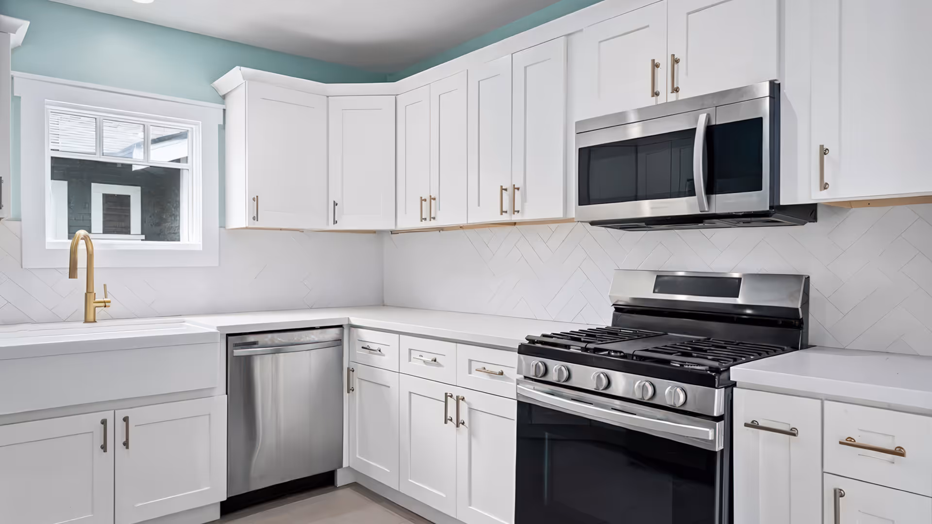 Modern kitchen with white cabinets, stainless steel dishwasher, stove, microwave, and gold faucet under a window.