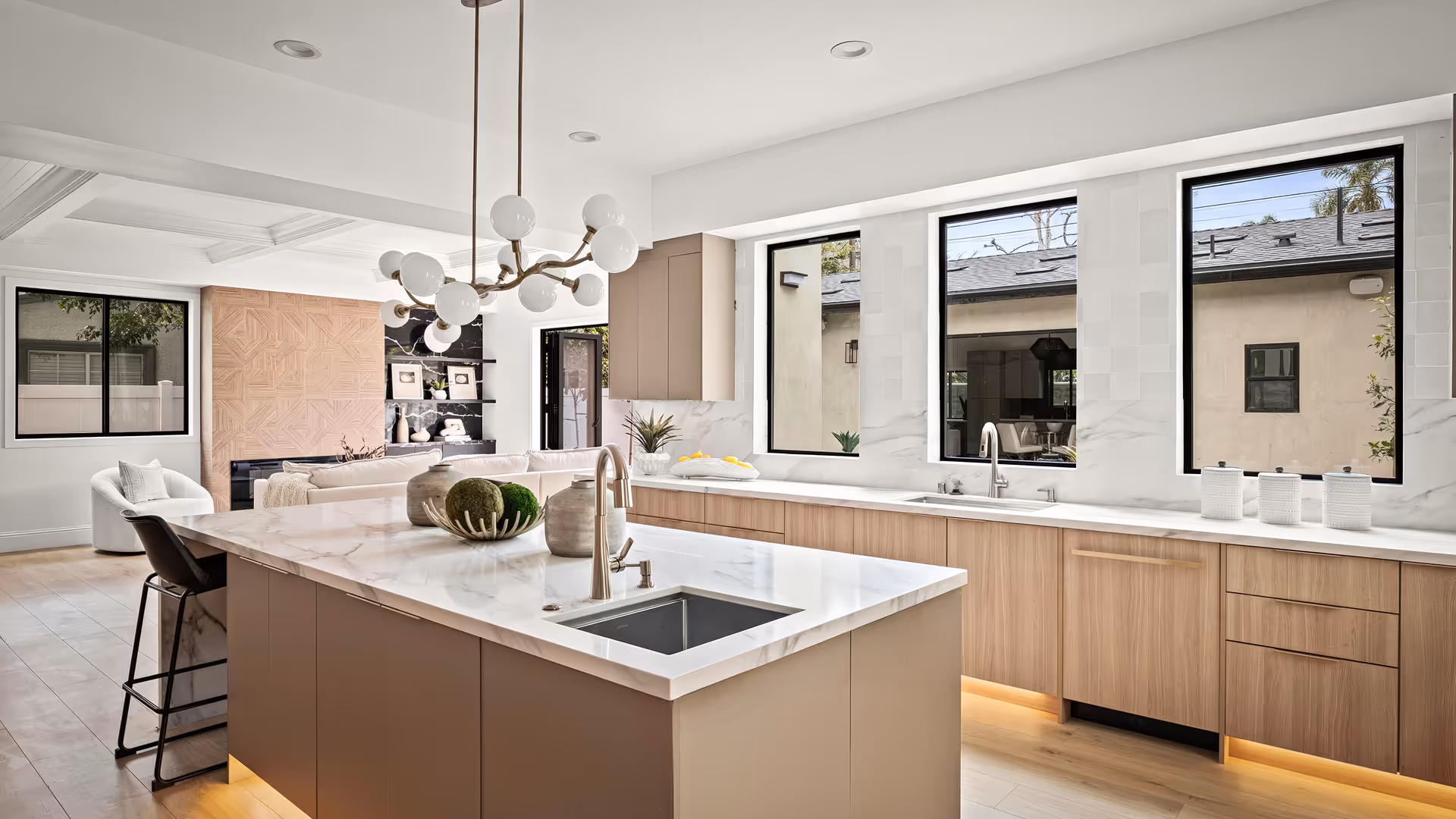 Modern kitchen with beige cabinets, white marble countertops, two sinks, large island, and three tall windows.
