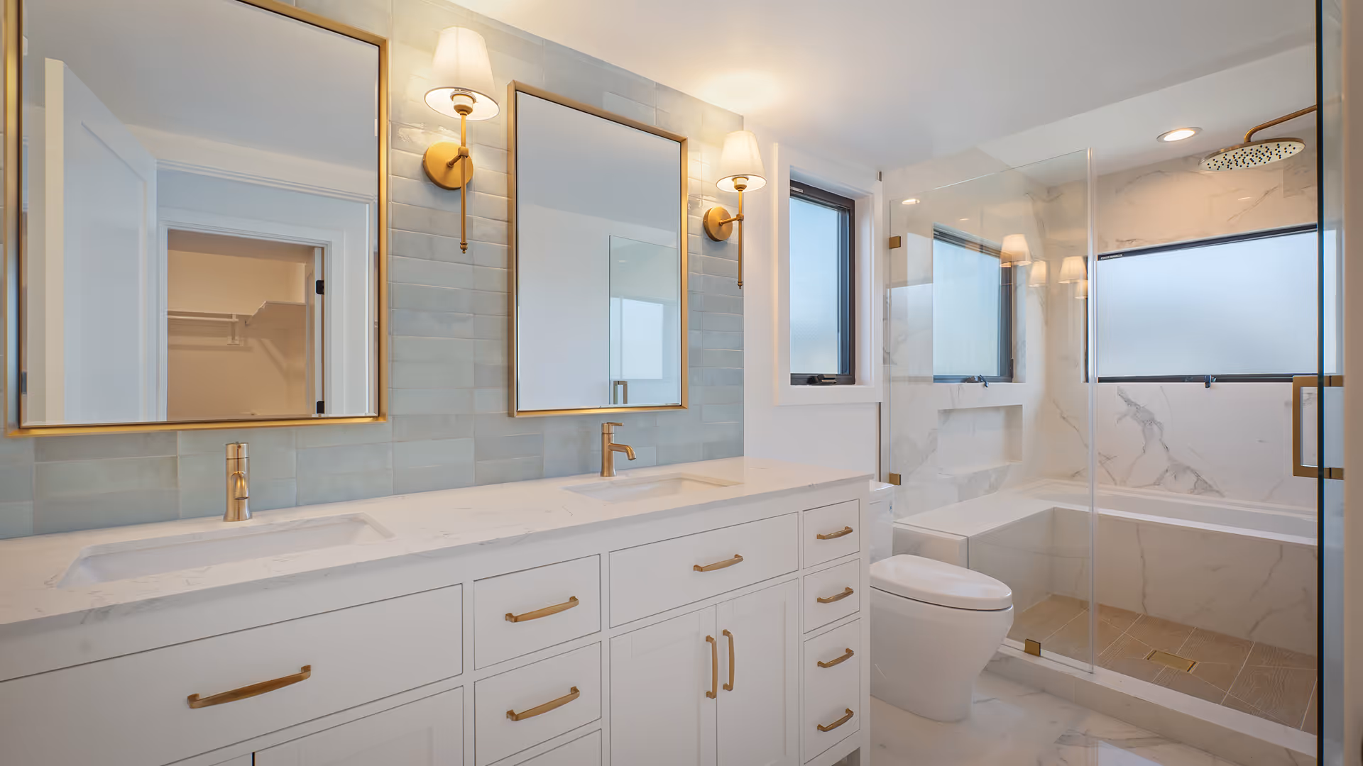 Modern bathroom with white double vanity, gold fixtures, two mirrors with wall sconces, and glass-enclosed shower with marble walls.
