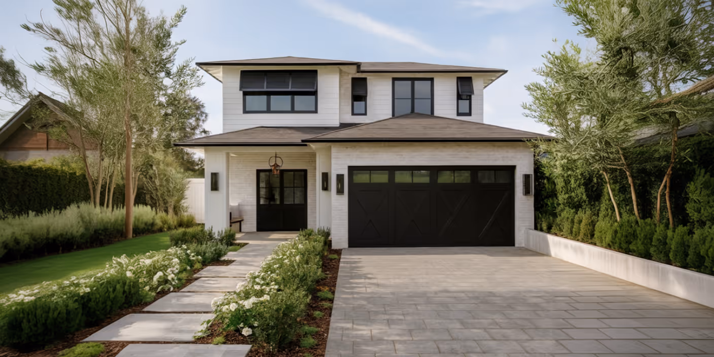 Modern two-story house with white exterior, black-framed windows and doors, and a large black garage door, surrounded by green landscaping and a paved driveway.