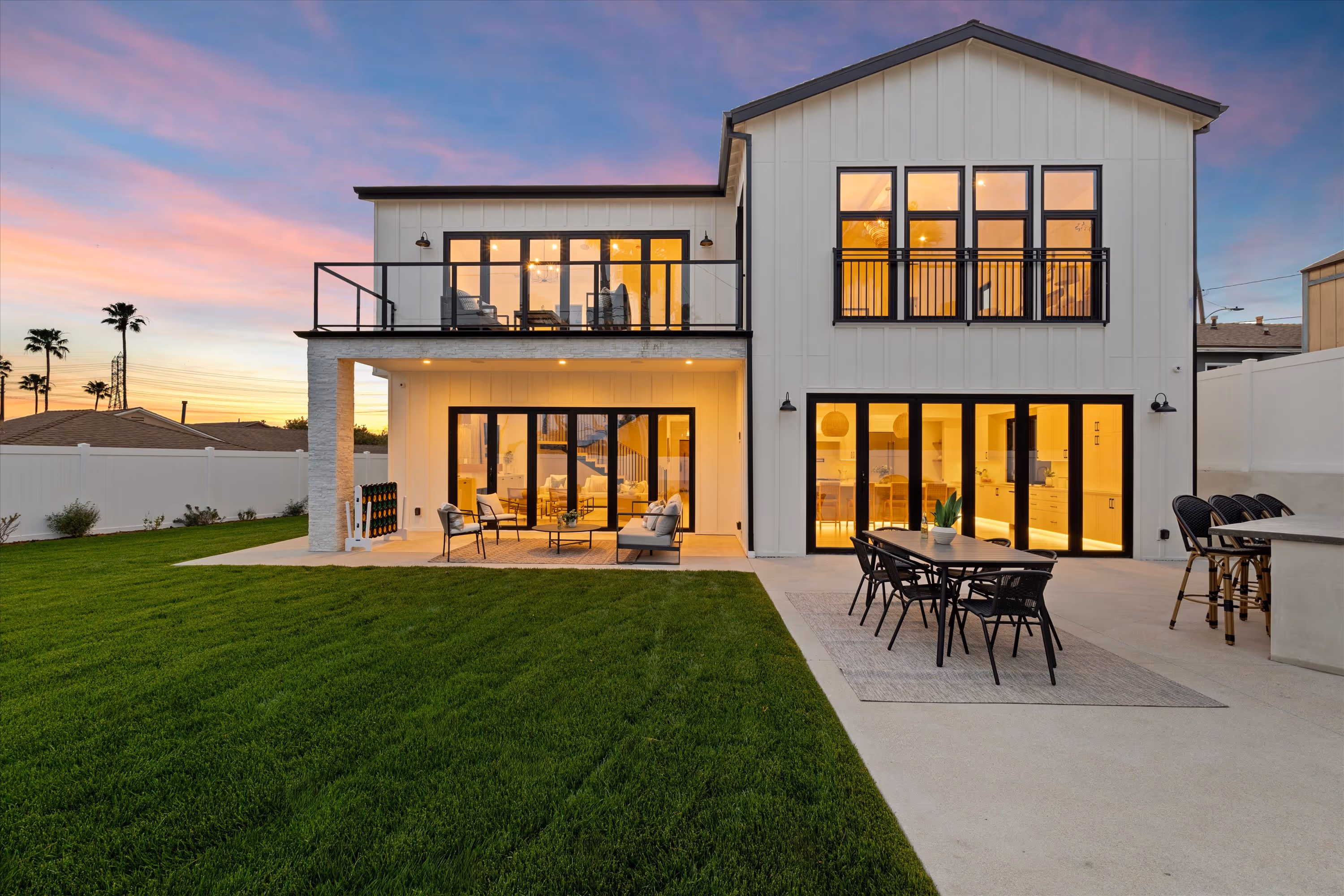 Modern two-story white house with large glass doors and windows, outdoor seating area, dining table, and green lawn at sunset.