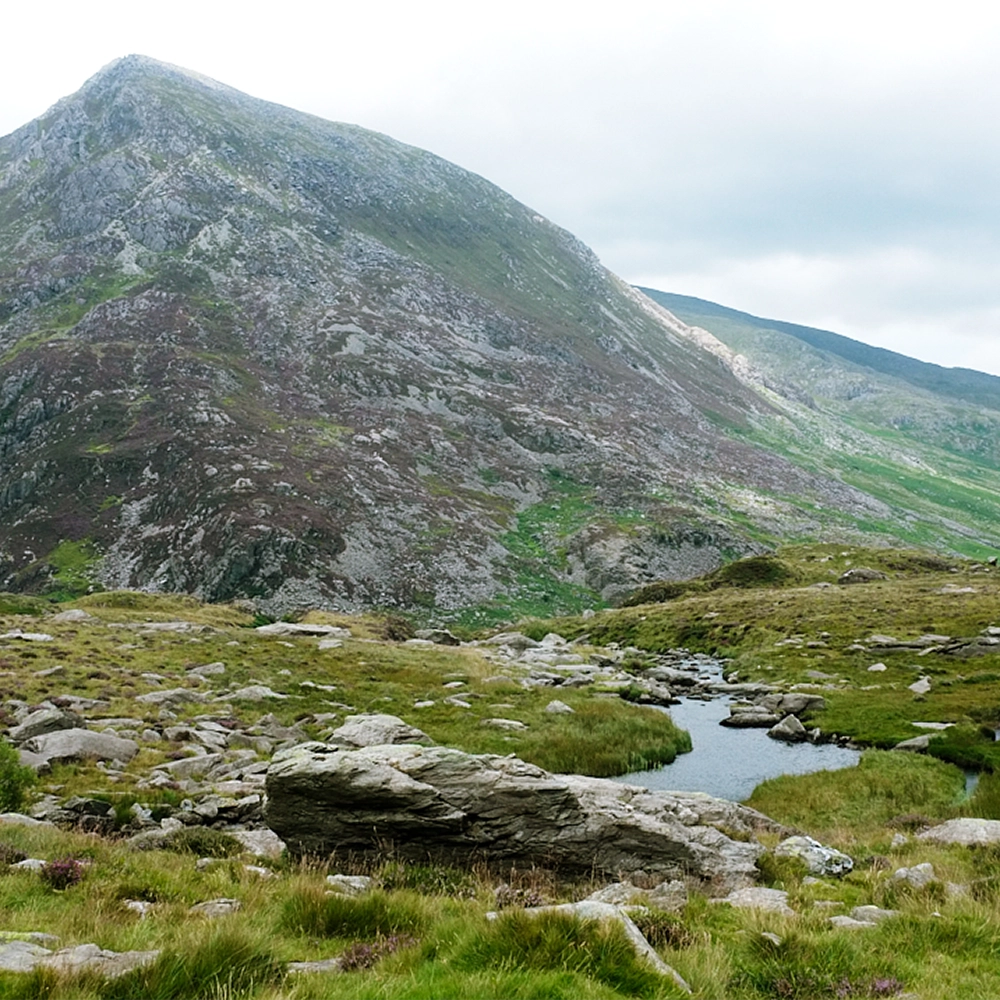 Rocky green mountain with patches of grass and a small stream running through grassy terrain in the foreground under a cloudy sky.