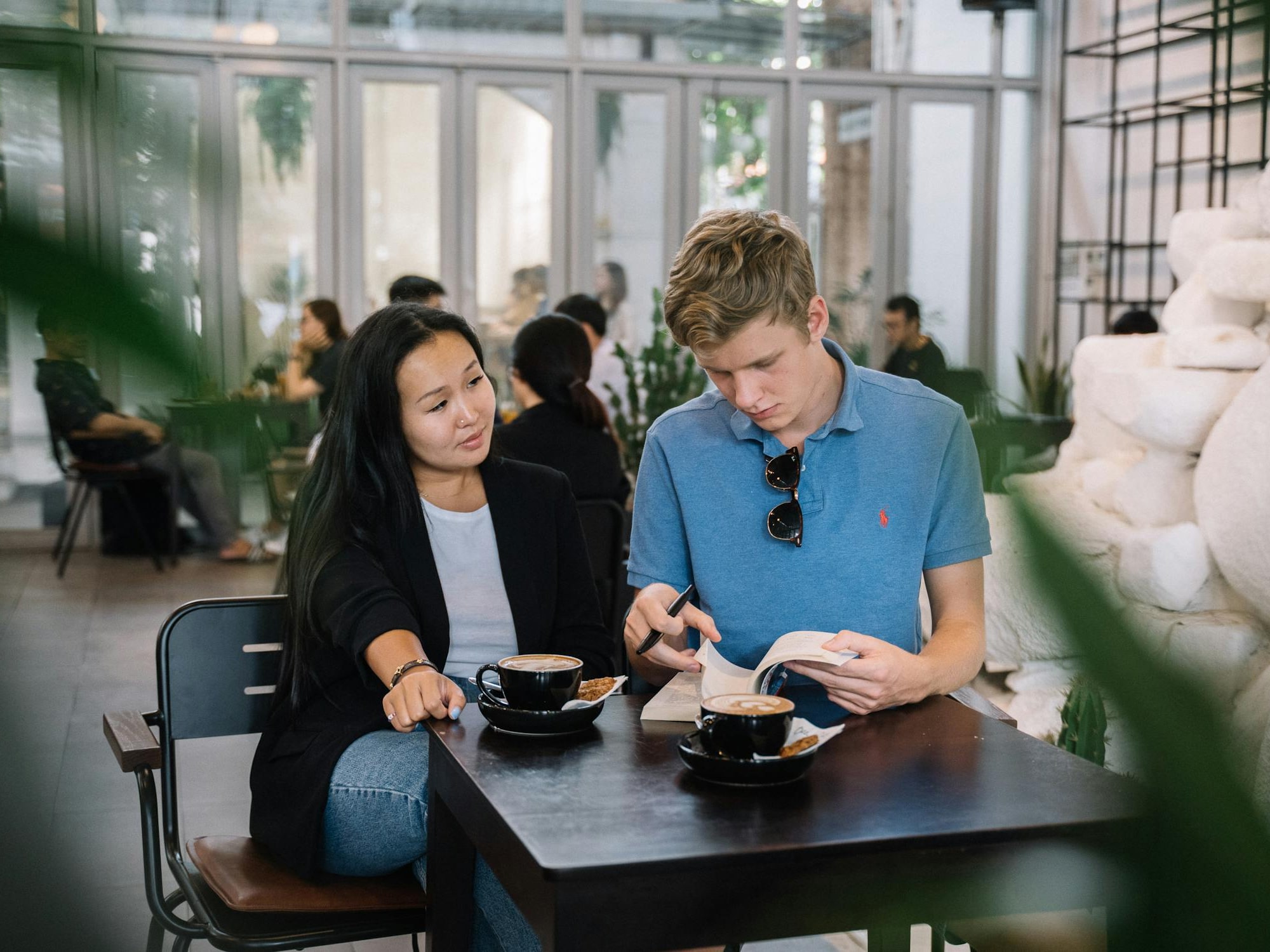 A young man and woman sit at a café table with two cups of coffee; the man reads a book while the woman looks on.
