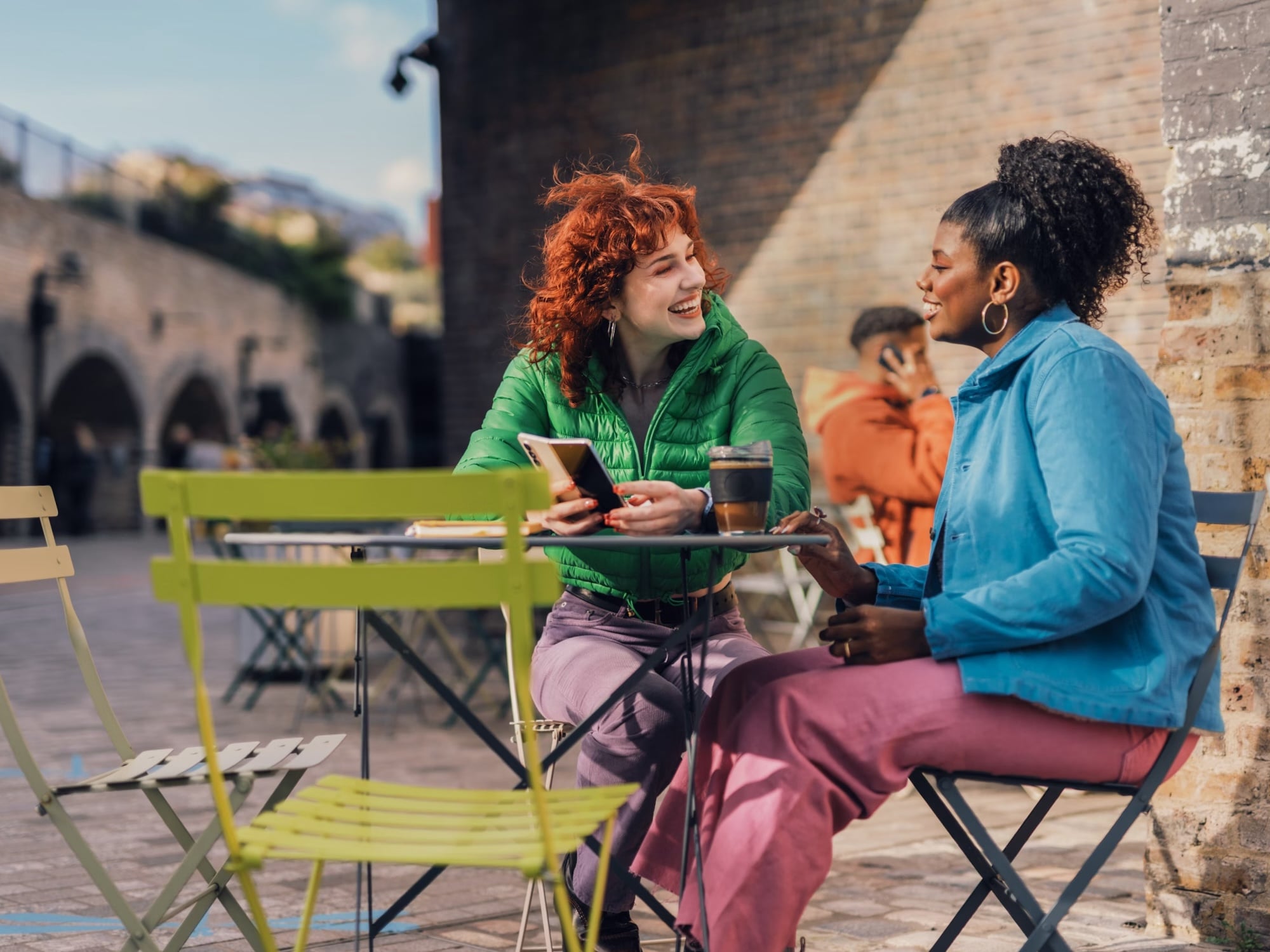 Two women sitting at an outdoor café table, smiling and chatting, one holding a smartphone and a cup of coffee nearby.