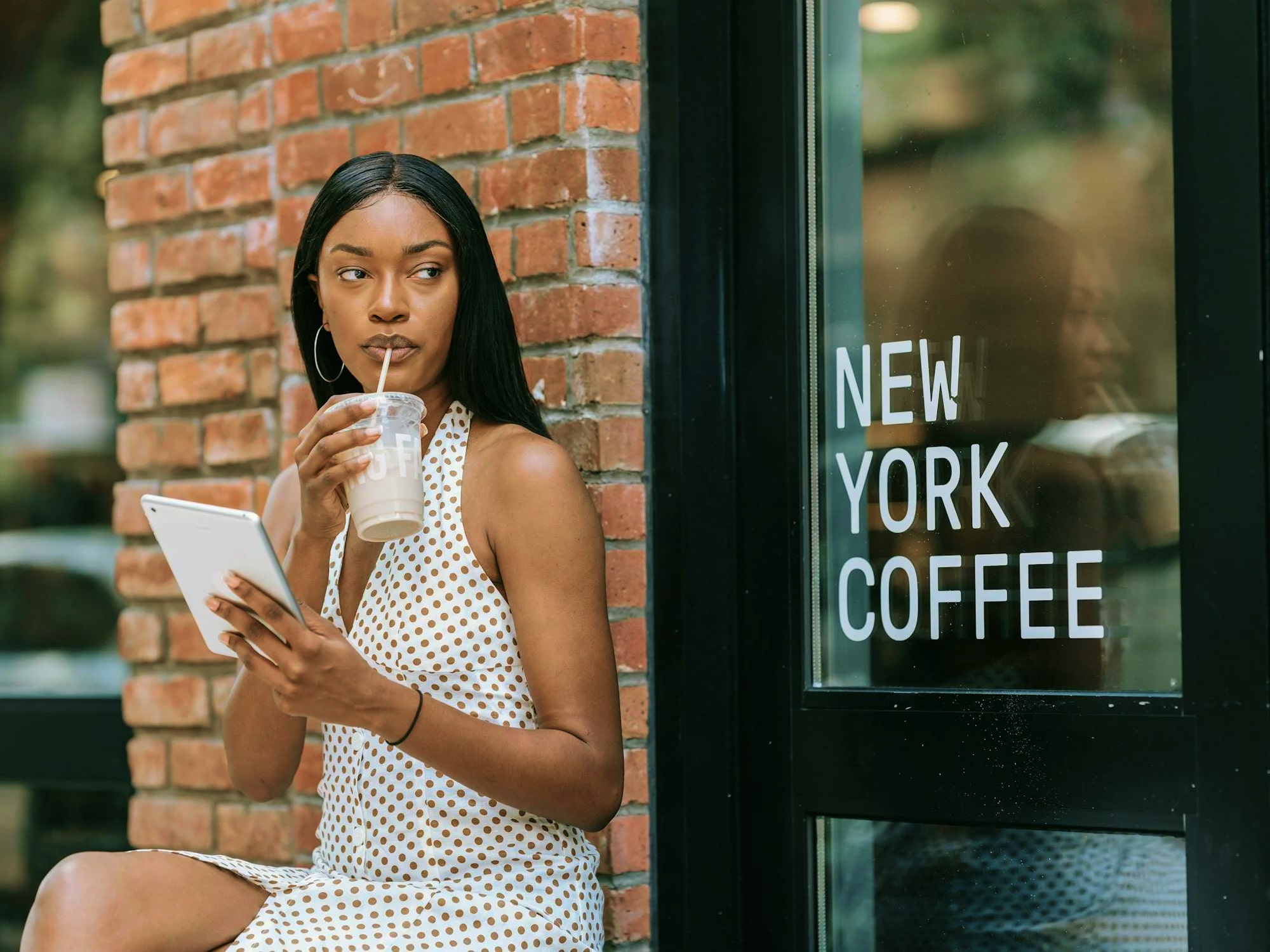 Woman in a white and brown polka dot dress sitting against a brick wall, drinking iced coffee and holding a tablet outside a cafe with a 'New York Coffee' sign on the window.