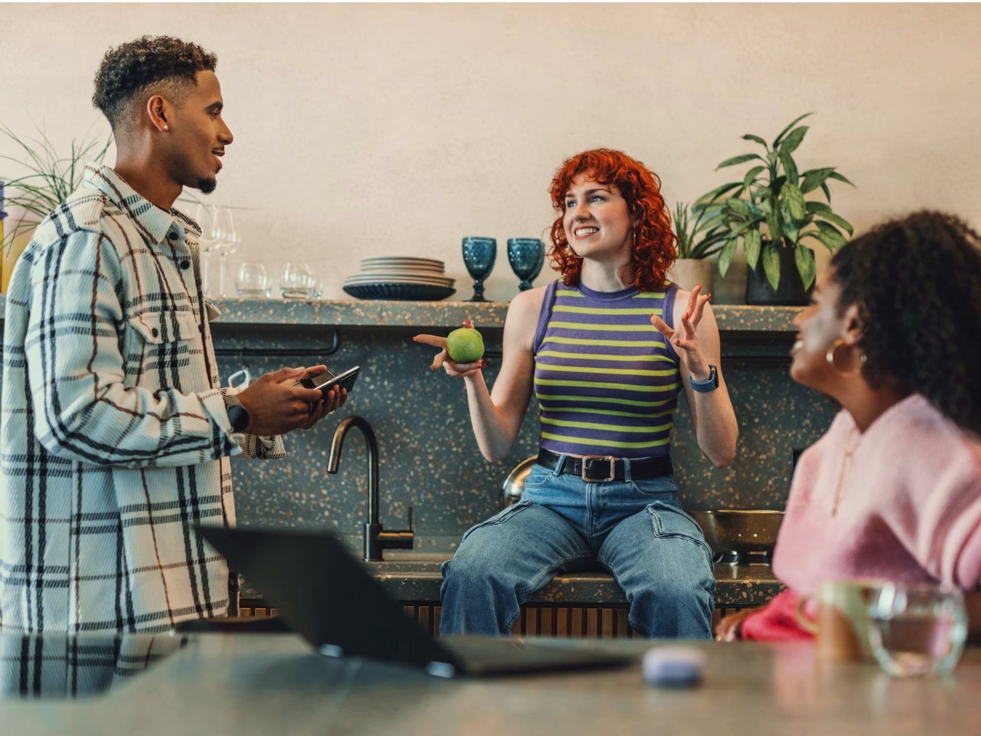 Three young adults having a lively conversation in a kitchen, with one holding a green apple and another holding a smartphone.