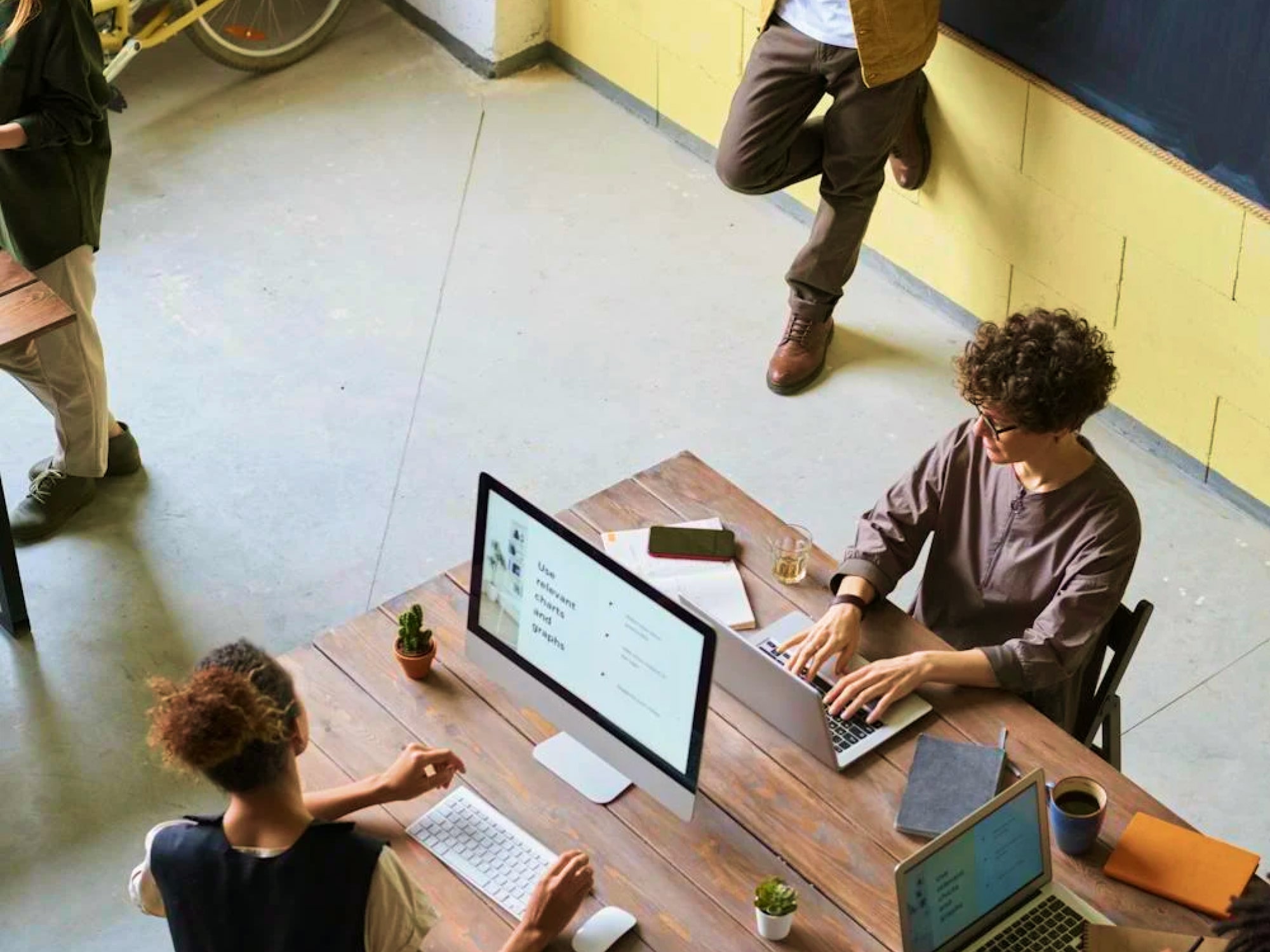 Two people working on laptops and a desktop computer at a wooden table in a bright office space.