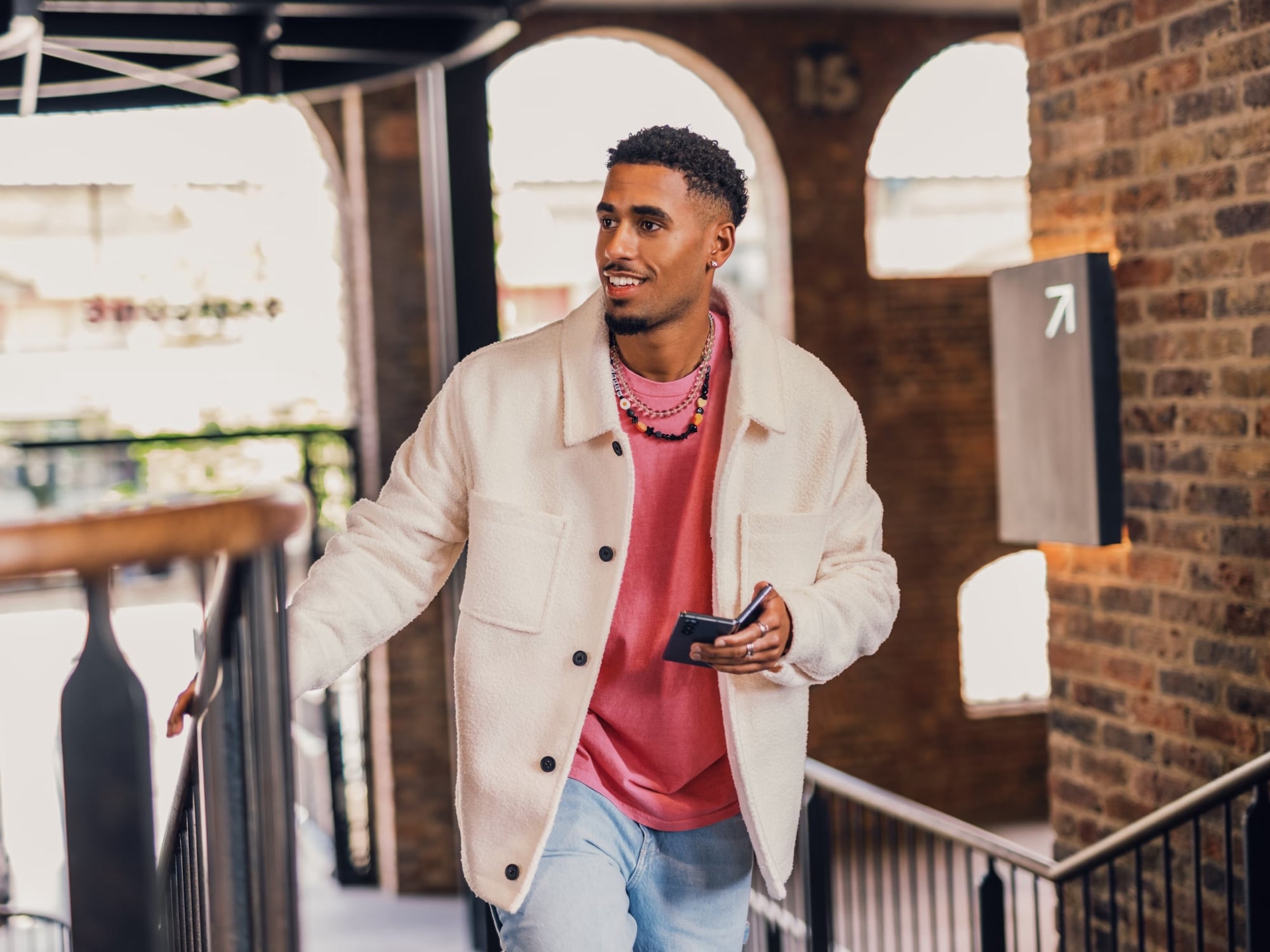Smiling young man in a white jacket and pink shirt holding a smartphone, walking up stairs in a brick interior.