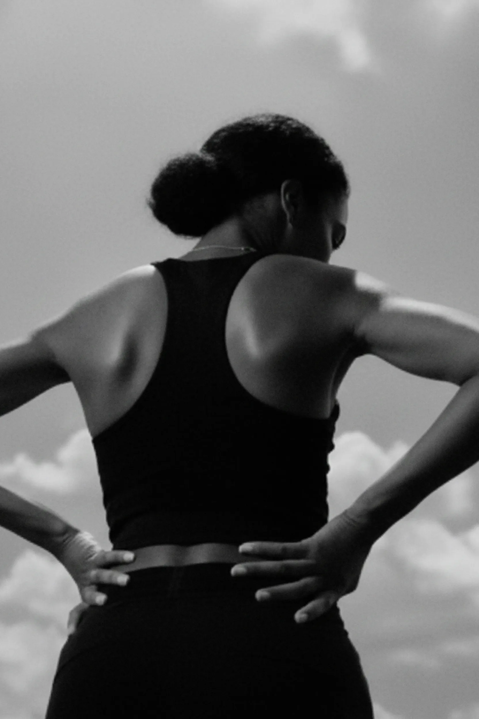 Black-and-white photo of a woman from behind with hair in a bun wearing a sleeveless top and placing hands on her hips against a cloudy sky.