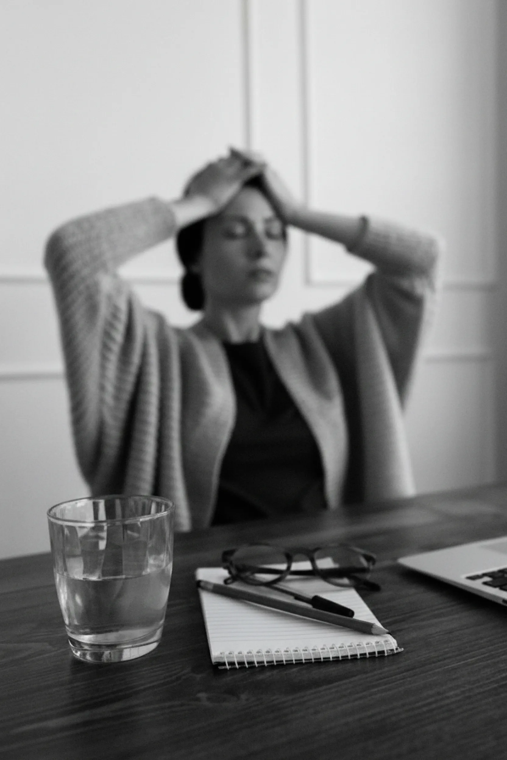 Woman sitting at a desk with hands on her head, with a glass of water, notebook, pen, and glasses in the foreground.