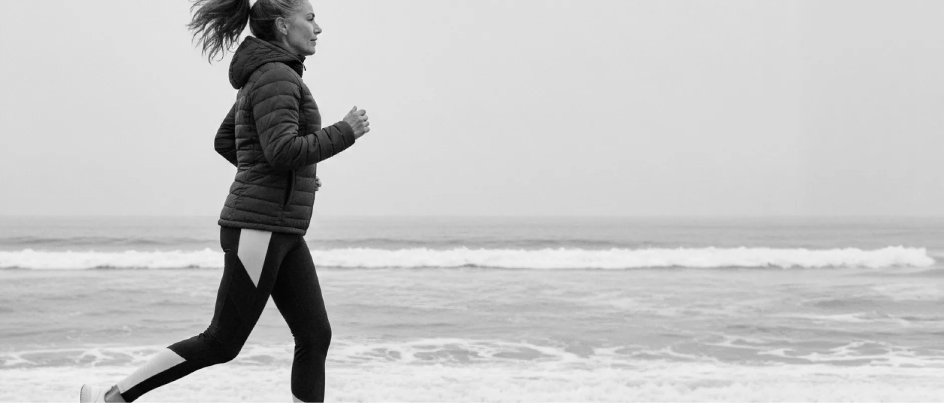 Woman jogging along the beach wearing a hooded jacket and leggings with ocean waves in the background.
