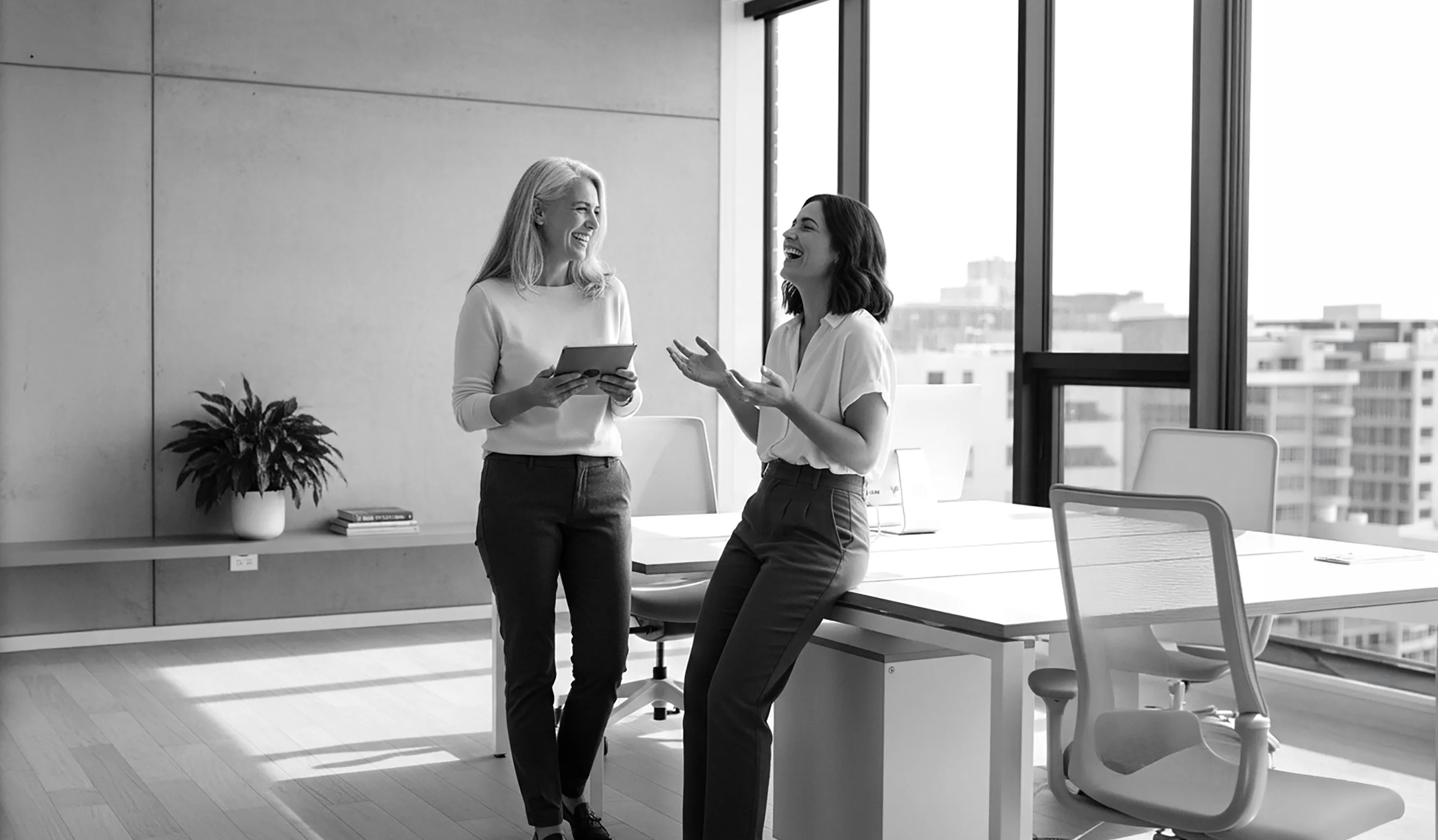 Two women smiling and talking in a modern office near a window with city view, one standing with a tablet and the other sitting on a desk.