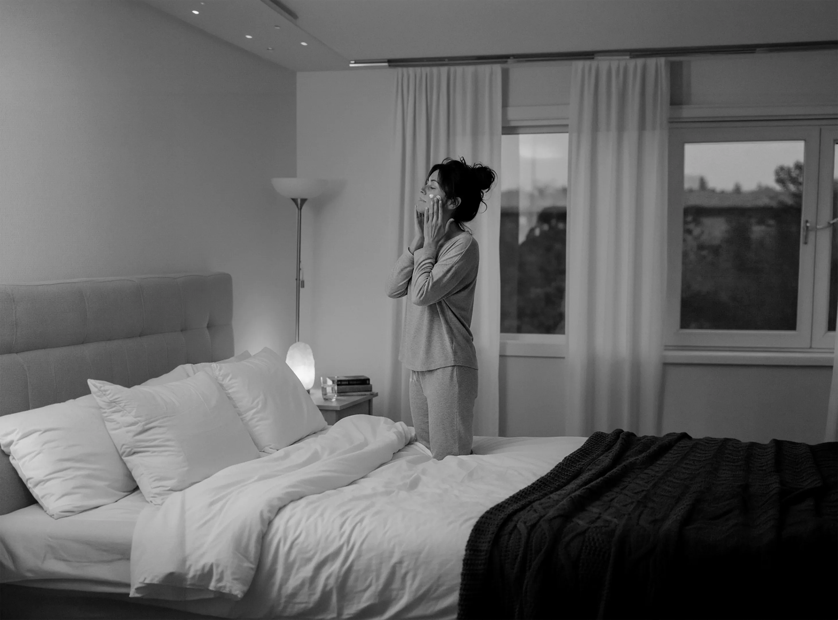 Woman in pajamas applying skincare cream to her face while kneeling on a bed in a softly lit bedroom.