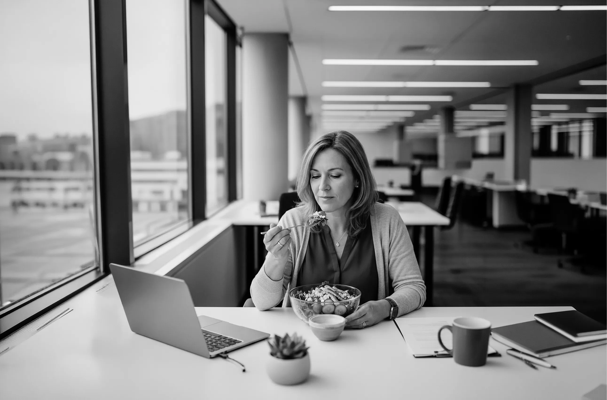 Woman eating salad at desk with open laptop, coffee, and notebooks in modern office.