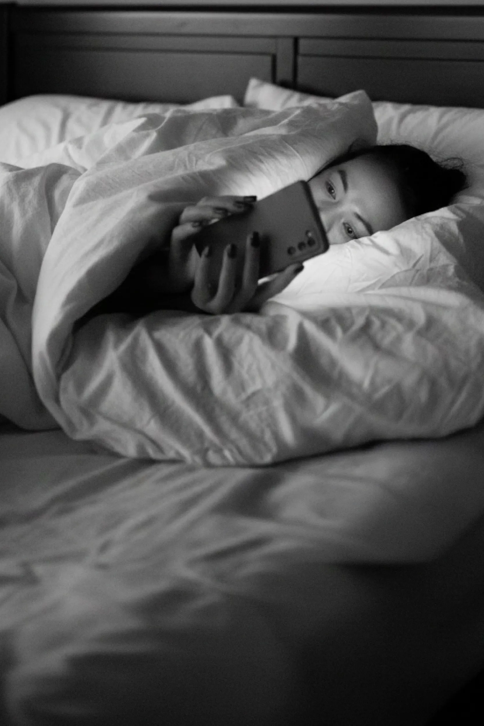 Person lying in bed under white blankets, looking at a smartphone.
