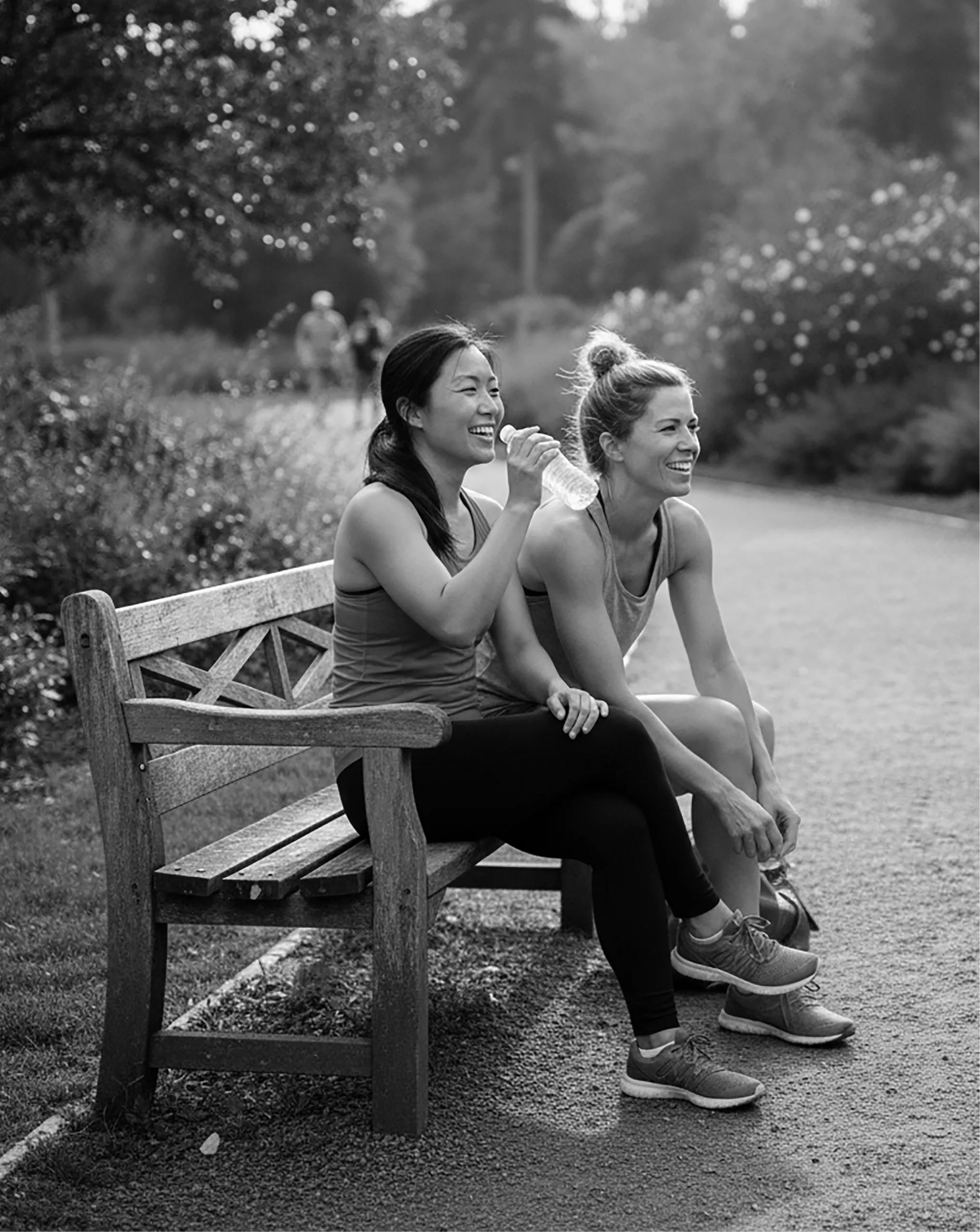 Two women in athletic wear sitting on a wooden bench outdoors, smiling and relaxing after exercise.