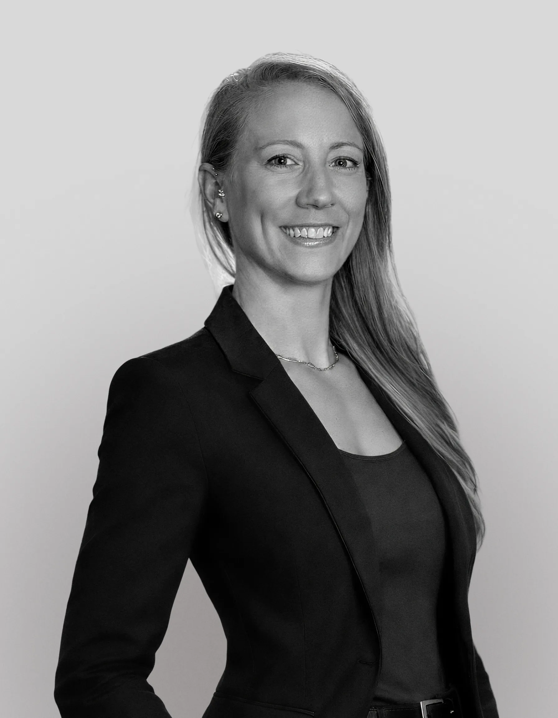 Smiling woman with long hair wearing a black blazer over a tank top, posed against a light gray background.